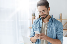 Glad Fashionable Young Bearded Guy Wears Stylish Clothes And Denim Shirt, Has Trendy Hairstyle, Happy To Exchange Messages With Friends, Uses Free Internet Connection On Electronic Modern Gadget