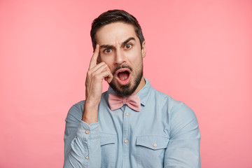 Wall Mural - People, facial expressions and emotions concept. Shocked amazed young male with trendy hairdo, wears formal clothes, keeps index finger on temple, keeps mouth widely opened, being emotional.