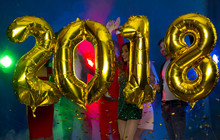The Composition Of The Golden Balloons In The Form Of Figures On A Background Of A Dancing Group Of Young People. The New Year 2018. Festive New Year Party Or Disco.