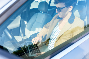 Wall Mural - Portrait of successful modern entrepreneur wearing sunglasses working, using laptop sitting in luxury car, view from behind glass window