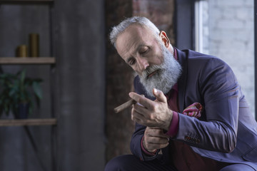 Wall Mural - Portrait of serious unshaven pensioner buttoning cuff link on sleeve of elegant shirt while holding cigar in arm. Relax concept