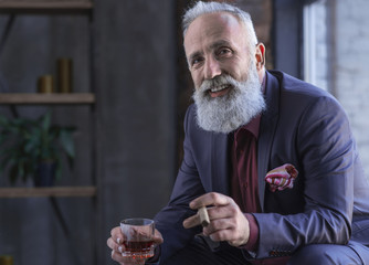 Wall Mural - Portrait of cheerful bearded old career man tasting cognac and keeping tobacco while locating in room. Rest concept