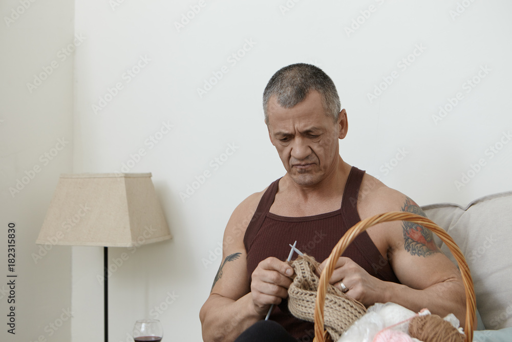 People, age, hobby and leisure concept. Portrait of concentrated elderly Caucasian man with grey hair and muscular arms with tattoo on shoulders focused on knitting, holding needles and brown wool - obrazy, fototapety, plakaty 