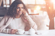 © Viacheslav Yakobchuk - Dreamy mood. Pretty black woman with curly hair keeping smile on her face holding white cup in right hand while sitting at the table