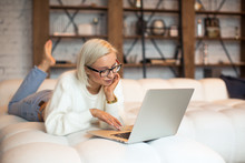 Pretty Young Blonde Girl Working On A Laptop Sitting On The Bed. Beautiful Young Woman In A Fluffy White Sweater And Glasses Working From Home On A Laptop Lying On The Bed In The Room With White Walls