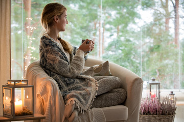 Young beautiful woman sitting home in the chair by the window with cup of hot coffee wearing knitted warm sweater. Cozy room decorated with lanterns and candles. 