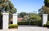 Fototapeta  - Black metal driveway entrance gates set in brick fence with garden trees in background