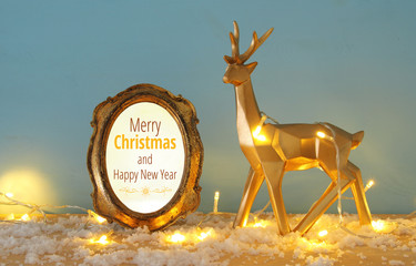 Gold shiny reindeer on snowy wooden table with christmas garland lights.