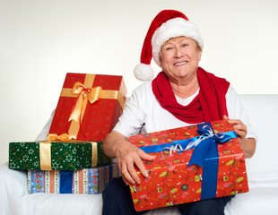 Wall Mural - Aged woman dressed in santa hat with gift boxes sit on sofa, white background. New year eve and christmas holiday concept.