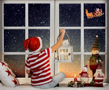 Merry Christmas And Happy Holidays!  Little Boy Sitting  On The Window And Looking At Santa Claus Flying In His Sleigh Against Moon Sky.  Room Decorated On Christmas.
