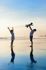 Wall Mural - Father tossing high in air baby son, mother jumping by water pool. Happy family walk with fun by sunset black sand beach with sea surf. Active parents, outdoor activity on summer vacation with kids.