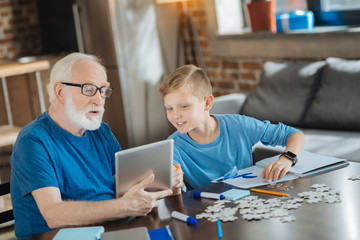 Wall Mural - Digital device. Joyful nice happy boy pointing at the tablet and smiling while helping his grandfather to use it