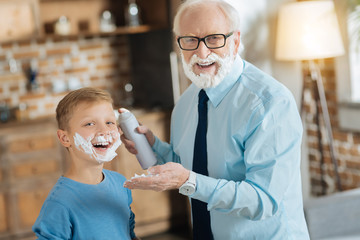 Wall Mural - Time for shaving. Happy nice elderly man holding shaving foam and smiling to you while standing together with his grandson