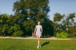 © ViDi Studio - Young pretty girl with long brown hair dressed in light clothes staying on green lawn grass in the park on trees background. Summer sunny time.