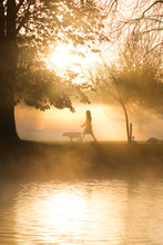 Woman Walking Dog In Early Morning Mist In Autumn Fall With Sunlight In Background