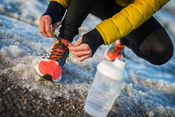 Close up of woman's hands tying shoelaces on snowy winter road with a bottle of water in front.