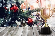 Champion Golden Trophy On Wooden Desk And Lens Flare Effect With  Decoration Ball Hanging On Christmas Tree With Snow Effect Background, Winner, Success And Congratulation Concept, Copy Space