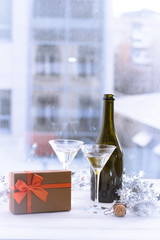 A romantic festive afternoon with two glasses chapmagne, a half full bottle, a brown gift box with an orange bow. A siver shiny garland on a white wooden table. A blurred window view at the background