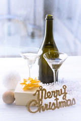 A romantic festive afternoon with two glasses and a bottle of chapmagne, a beige gift box with a golden bow. A silver garland, white and gold Christmas tree balls and gold Merry Christmas inscription