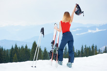 Rear View Of Active Female Skier Wearing Ski Pants And Red Bodice, Holding Helmet Above A Head, Standing On The Top Of The Snowy Hill With Ski Equipment At Winter Ski Resort In The Mountains Copyspace