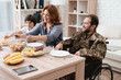 A veteran in a wheelchair dines with his family. A man in uniform is sitting at the kitchen table