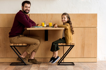 smiling daughter and father sitting at table in cafe and looking at camera