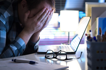 Problems at work. Sad, unhappy and tired man covering face with hands in office or home at night. Burnout, stress, workplace bullying or depression concept. Glasses on table.