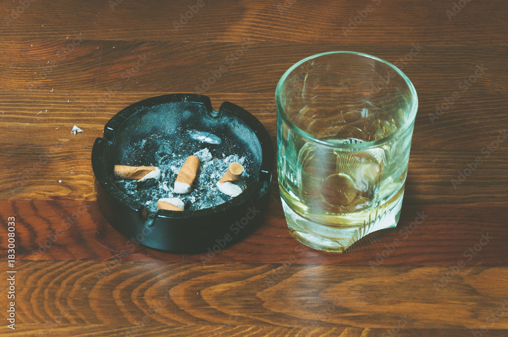 Glass with alcohol drink and dirty black ceramic ashtray full of cigarette  butts on the wooden table surface close up selective focus Stock Photo