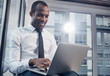 © Yakobchuk Olena - Daily life. Low angle of delightful stylish young african businessman is smiling while sitting on windowsill and typing text on laptop. Window in background with copy space in the right side