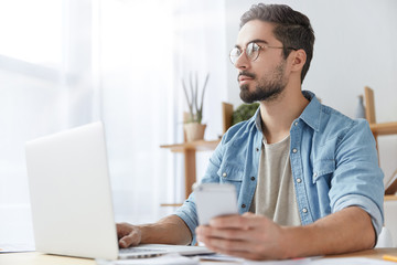 Thoughtful beared male student in stylish clothes surrounded with modern gadgets, being always in touch, uses free internet connection for searching information to prepare sceintific report.