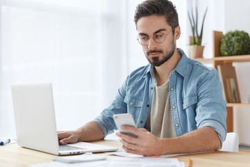 Stylish bearded male freelancer works remotely at home, uses modern electronic devices and internet connection, being always in touch, recieves messages and keyboards on computer. Technology, people