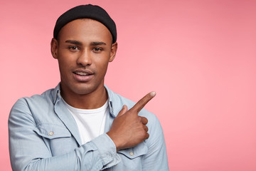 Attractive young male has specific appearance, wears trendy black hat and denim shirt, points with fore finger at copy space of pink background. Look at this! People and advertising concept.