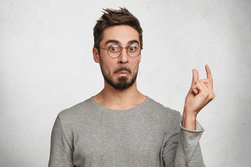 Wall Mural - Surprised bearded young male with trendy hairdo shows something little or tiny with fingers, wears casual sweater, isolated over white background. Shocked fashionable guy demonstrates something