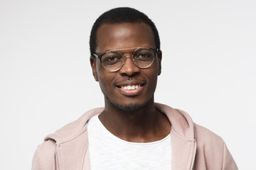 Wall Mural - Indoor portrait of young handsome African male pictured isolated on white background wearing white T-shirt, pink hoodie and round eyeglasses showing relaxed smile and looking confident and satisfied