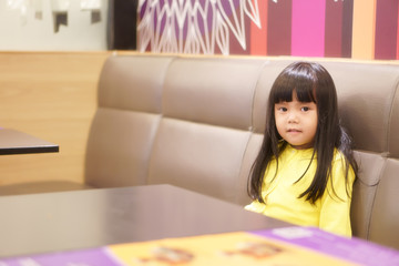 Wall Mural - Asian children cute or kid girl relax sitting for waiting order or food for lunch or dinner on long sofa and wood table at cafe restaurant with wear yellow shirt and copy space