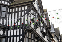 Old Arts And Crafts Buildings With Traditional Black And White Ornaments.. Road Is Decorated With Green And Red Baubles And Lights For Christmas Celebration. Overcast Sky.
