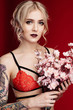 Fashion style, model test. Good for lingerie shops and tattoo content. Beautiful blond woman with tattoo in a sexy red lingerie on a red background. Sakura flowers in hands