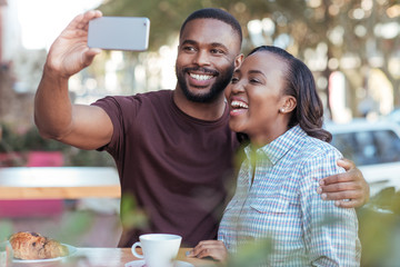 Wall Mural - Laughing young African couple taking selfies at a sidewalk cafe