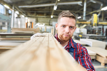 Wall Mural - Head and shoulders portrait of handsome young factory worker looking at camera while carrying long wooden board moving material in workshop, copy space