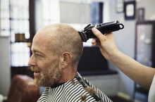 A Customer Sitting In The Barber's Chair, And A Barber Using An Electric Shaver To Shave His Head. 