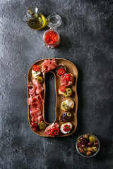 Wall Mural - Tapas or bruschetta variety. Crusty bread with ham prosciutto, sun dried tomatoes, olive oil, olives, pepper on decorative wooden plate over dark texture background. Top view with space