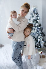 Happy woman,cheerful mother and little children,a girl and a newborn boy sitting on a fluffy white blanket beside the Christmas festive green Christmas tree with white toys,snowflakes,glowing lights