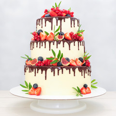 Wall Mural - Three-tiered wedding cake in chocolate, decorated with slices strawberries, blueberries, figs, red currants and green leaves on a white background. Picture for a menu or a confectionery catalog.