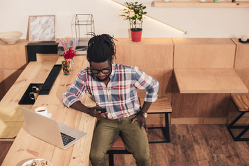 african american man using laptop and smartphone at bar counter in cafe