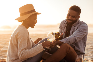 Wall Mural - Smiling African couple drinking wine at the beach at sunset