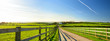 Fence casting shadows on a road leading to small house between scenic Cornish fields under blue sky, Cornwall, England