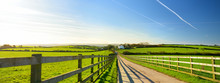 Fence Casting Shadows On A Road Leading To Small House Between Scenic Cornish Fields Under Blue Sky, Cornwall, England