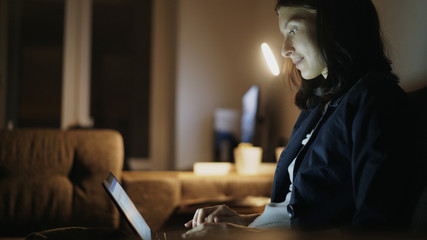 Young pretty woman using laptop computer and surfing social media sitting on coach at home in night time