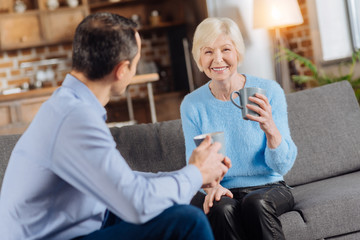 Wall Mural - Pleasant conversation. Cheerful senior woman and her young son sitting in the living room, drinking coffee and talking to each other