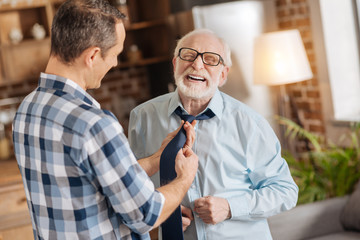 Sonly care. Pleasant young man tying the tie of his elderly father while the man laughing, closing his eyes